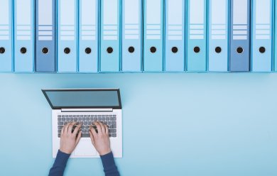 Archiv mit blauen Ringordern und eine Person, die am Laptop arbeitet